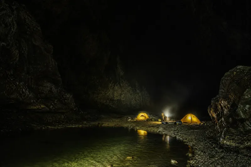 巨大洞﻿厅内两顶发光帐篷组成的地下营地​，队‍员在暗河​边生火﻿做饭﻿